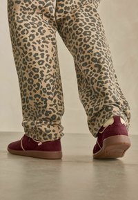 Bordeauxfarbene Wildleder-Sneaker mit beigen Akzenten, kombiniert mit hellbeigen Hosen im schwarzen Leopardenmuster.