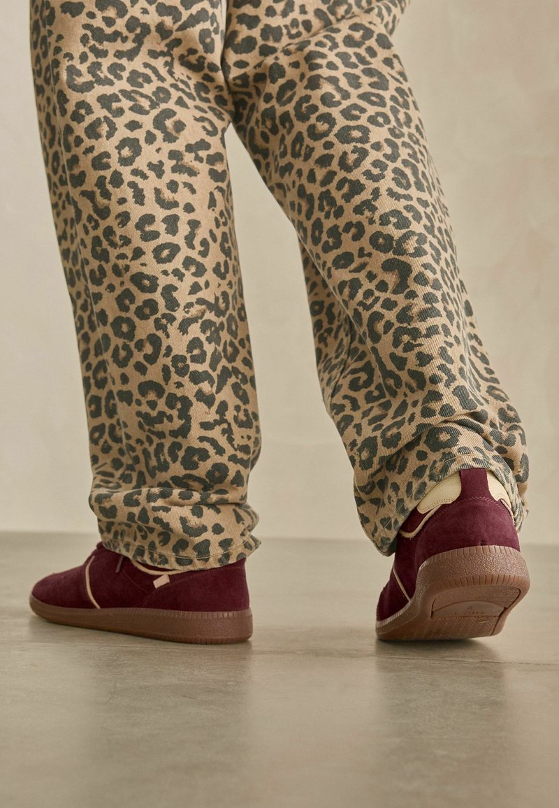 Bordeauxfarbene Wildleder-Sneaker mit beigen Akzenten, kombiniert mit hellbeigen Hosen im schwarzen Leopardenmuster.