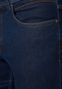 Granatowe dżinsy z pomarańczowymi przeszyciami, z przednią kieszenią i gładką fakturą. Zawierają metalowy detal z guzikem obok kieszeni.