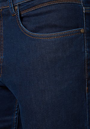 Granatowe dżinsy z pomarańczowymi przeszyciami, z przednią kieszenią i gładką fakturą. Zawierają metalowy detal z guzikem obok kieszeni.