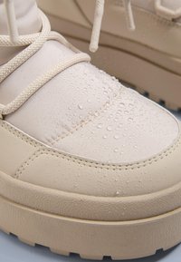 Lichtbeige waterdichte sneaker met een gestructureerd oppervlak en druppels, voorzien van een dikke buitenzool en witte veters. Rond, gewatteerd ontwerp.