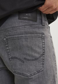 Grå denimjeans med en baklomme med buet søm og et svart lærmerke som viser merkeinformasjon og detaljer om håndverk.