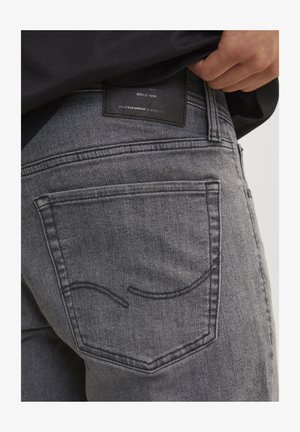 Pilkos džinsinio audinio kelnės su užpakaliniu kišeniu, turinčiu kreivą siuvinėjimą, ir juodos odos etikete, kurioje pateikiama prekės ženklo informacija ir meistriškumo detalės.