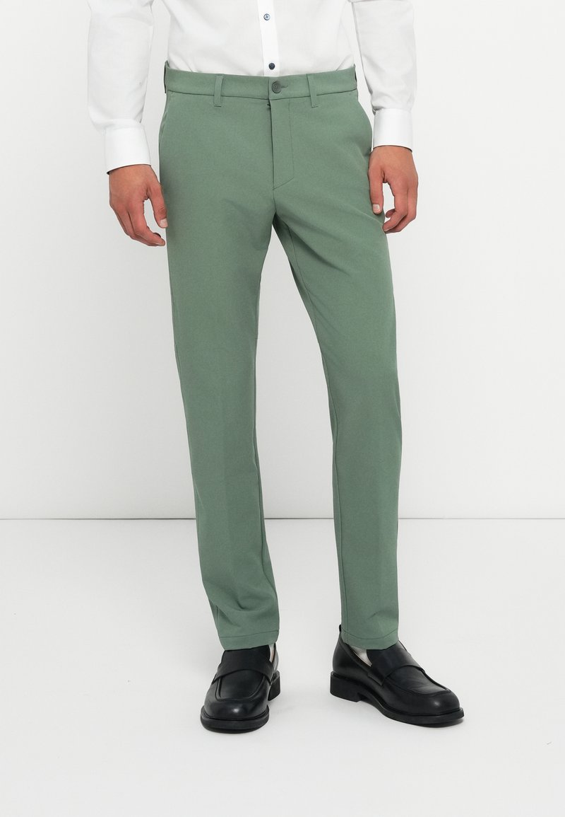 Green tailored pants made of smooth fabric, featuring a straight leg design, side pockets, and a button closure. Worn with black shoes.