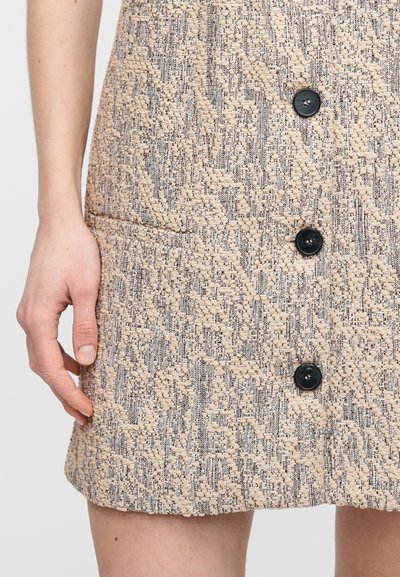 Φούστα από μπεζ υφή υφάσματος με υφαντό σχέδιο, που διαθέτει τρία μαύρα κουμπιά και πλαϊνή τσέπη. Λεπτή, δομημένη σιλουέτα.