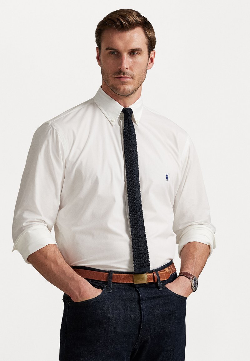Chemise blanche à manches retroussées, cravate noire texturée, ceinture en cuir marron et jeans foncés. Montre en accent au poignet. Coupe près du corps.