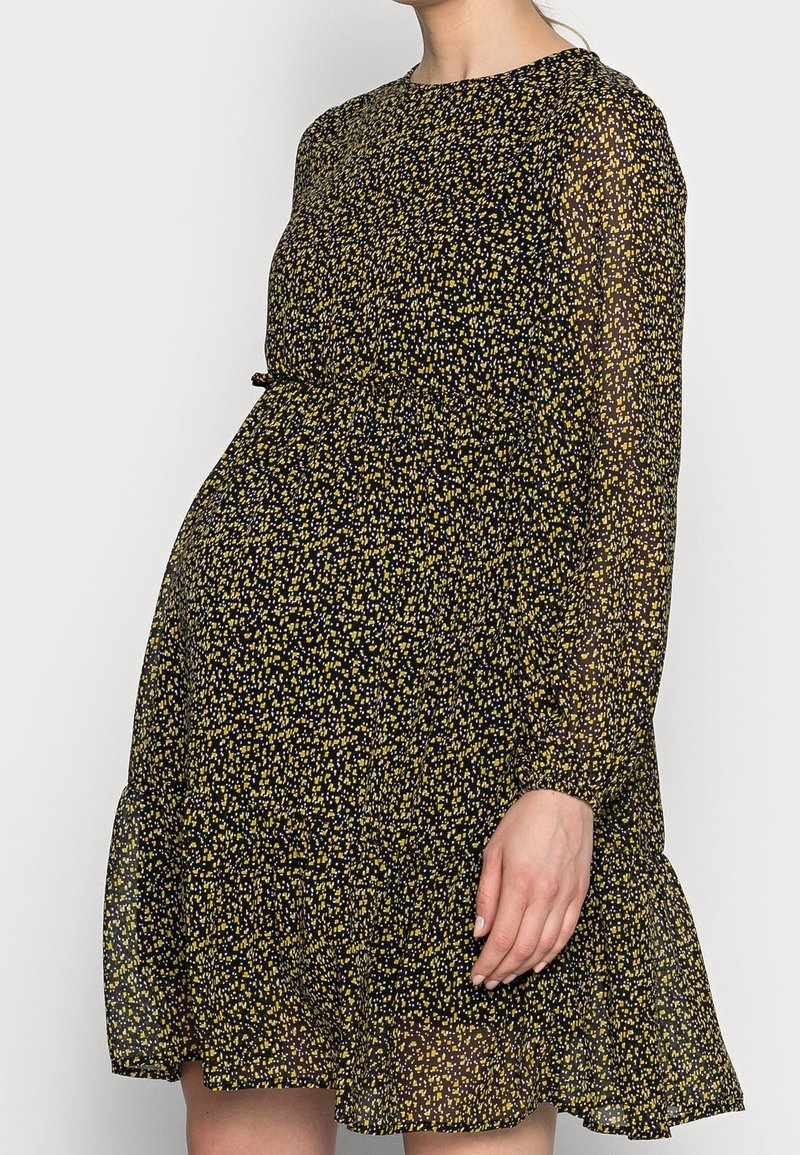 Femme portant une robe noire à manches longues avec un petit motif floral jaune et blanc, debout dans une posture détendue devant un fond uni.