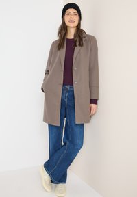 Ruskohvioinen villasekoitteen takki, jossa on kaksi nappia, yhdistetty tummansinisisiin leveisiin farkkuihin ja vaaleisiin lenkkareihin, neutraalin taustan edessä.