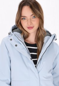 Jachetă impermeabilă de culoare albastră deschisă, cu fermoar pe față, glugă și detalii cu capse. Prezintă o căptușeală gri și o cămașă stripedă alb-negru dedesubt.