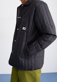 Czarna pikowana kurtka z zatrzaskami i kieszenią na piersi z logo. Zaokrąglony kołnierz i boczne kieszenie, w połączeniu z zielonymi spodniami.