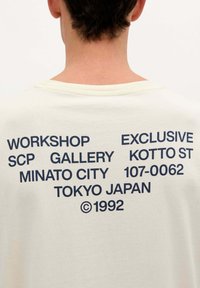 Tricou din bumbac crem cu text albastru marin pe spate: "WORKSHOP EXCLUSIVE SCP GALLERY KOTTO ST MINATO CITY 107-0062 TOKYO JAPAN ©1992".