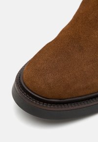 Botă din suede maro cu vârf rotunjit și talpă din cauciuc închis, având cusături contrastante pe margine. Textură netedă pe partea superioară.