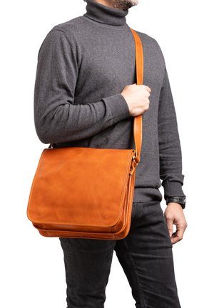 Tan leren messenger bag met een gladde textuur, opgevouwen flap en verstelbare schouderriem, gepresenteerd op een persoon die een grijze trui draagt.