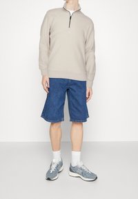 Persona che indossa un maglione beige a mezze zip, pantaloni shorts in denim blu fino al ginocchio, calze bianche e sneakers grigie New Balance, in piedi su un pavimento semplice.