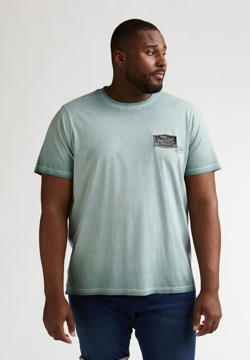 T-shirt en coton vert clair avec un col rond, des manches courtes et un patch noir avec un logo sur la poitrine gauche.