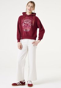Bordeaux hoodie met bergafbeelding en tekst, gecombineerd met witte wijde broek en rode sneakers met witte strepen. Eenvoudige gevlochten haarstijl.