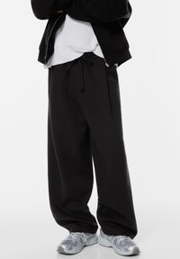 Schwarze weite Sweatpants mit einem Kordelzugbund, kombiniert mit einem weißen gerippten Shirt und silbernen Sportschuhen mit Mesh-Details.