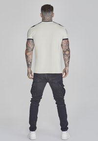 Camisa de corta manga de color claro con detalles en azul marino, usada con pantalones cargo negros y zapatillas blancas. Tatuajes visibles en los brazos.