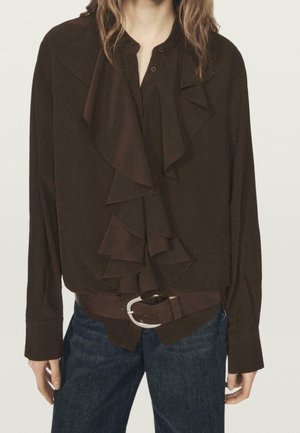 Kobieta ubrana w ciemnobrązową bluzkę z falbanami, wpuszczoną w niebieskie dżinsy z pasującym brązowym paskiem z srebrną klamrą.