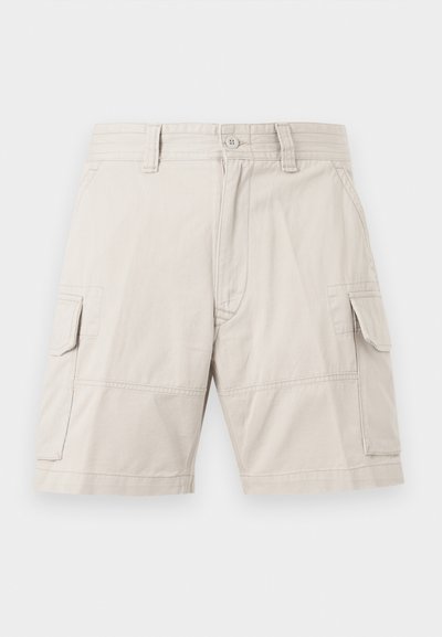 Μπεζ cargo σορτς με μπροστινό κουμπί και φερμουάρ, θηλιές για ζώνη, πλαϊνές τσέπες με καπάκι και μήκος μέχρι το γόνατο, εμφανιζόμενα σε απλό φόντο.
