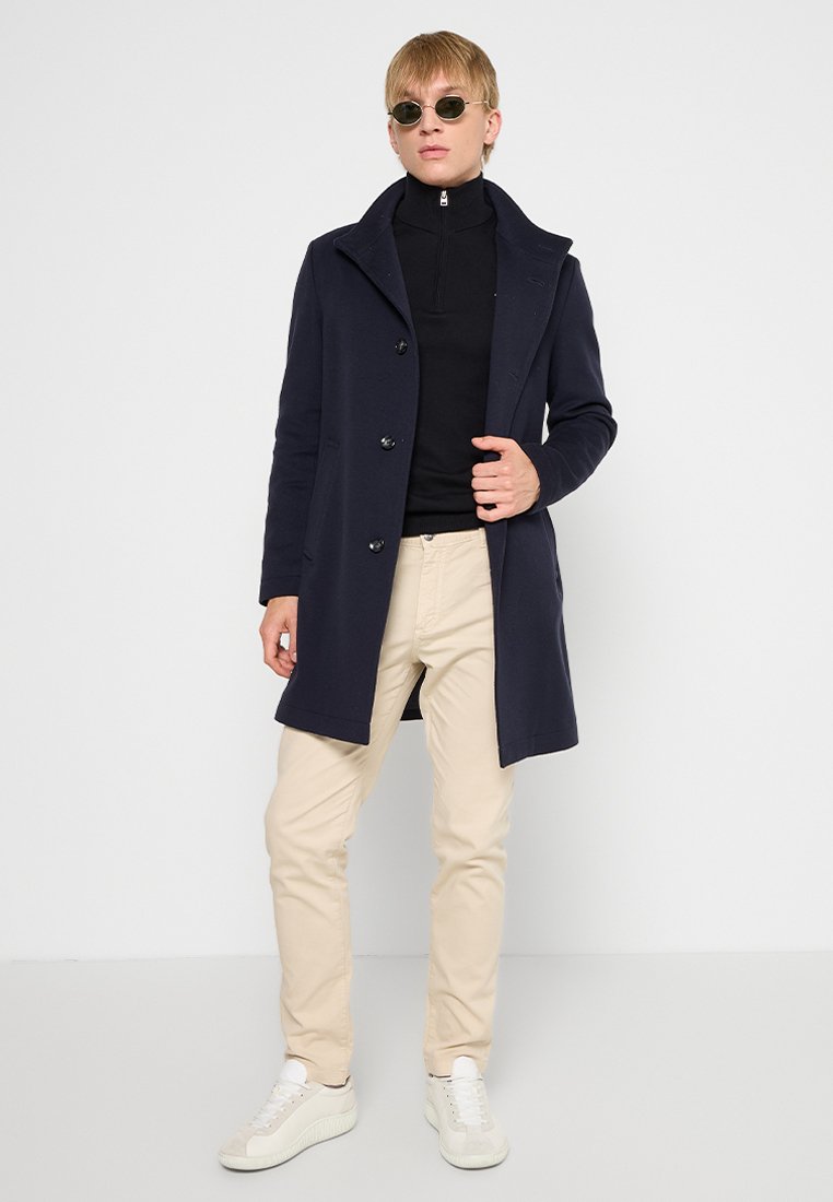 Manteau en mélange de laine bleu marine avec un large revers, six boutons et poches latérales, porté sur un col roulé noir et un pantalon beige avec des baskets blanches.