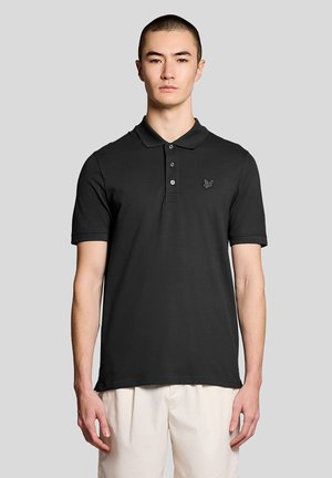 Mand iført en sort poloshirt med korte ærmer og en lille sommerfugl-logo på venstre bryst samt lysfarvede bukser.