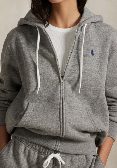 Γυναίκα που φοράει γκρι φούτερ με φερμουάρ και λευκά κορδόνια πάνω από λευκό ριμπ μπλουζάκι, σε συνδυασμό με matching γκρι φόρμα με λευκό κορδόνι.