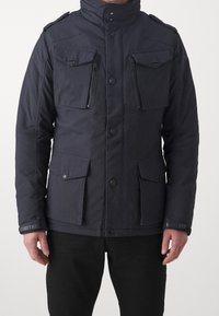 Granatowa ocieplana kurtka z czterema przednimi kieszeniami, zapinanymi na zamek kieszeniami na piersi oraz zapięciem na guziki. Posiada wysoki kołnierz i teksturowaną tkaninę.