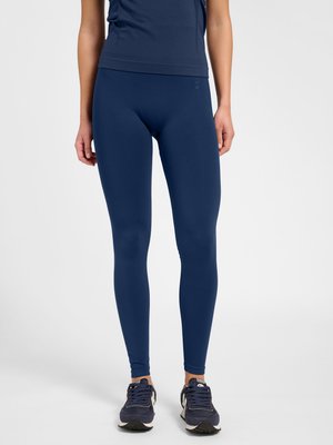 Kvinde iført marineblå tætsiddende leggings, matchende ærmeløs top og mørkeblå løbesko, stående på en ensartet lys baggrund.
