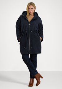 Quilted marineblå parka med hette, snøring i livet og frontlommer; har glidelåslukking og kontrasterende tekstur detaljer.