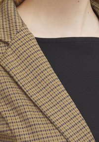 Revers de blazer en tartan beige avec des accents violets, associé à un haut noir côtelé. Mise en valeur des motifs et des textures des deux vêtements.