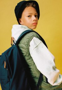 Gilet trapuntato verde sopra un hoodie bianco, cappello a secchio nero e zaino blu navy con dettagli in pelle marrone. Sfondo giallo.