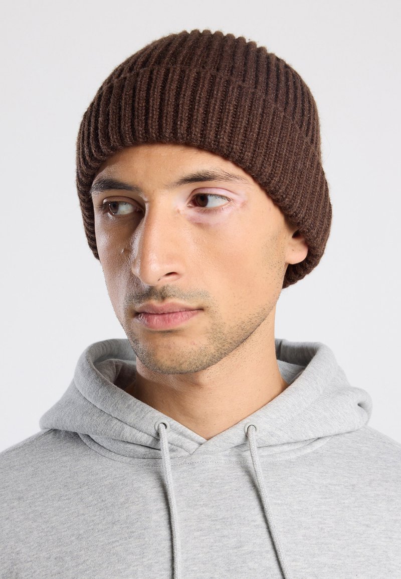 Beanie a coste marrone realizzato in lana, indossato aderente sulla testa. Abbinato a una felpa grigia chiara con un colletto a coulisse.