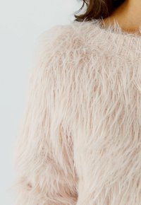 Close-up van een persoon die een zachte, pluizige, lichtroze trui draagt met een gestructureerd, verenachtig breipatroon.