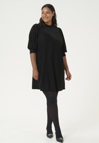 Robe noire plissée avec un col haut et des manches bouffantes. Portée sur des collants opaques noirs et assortie avec des chaussures à talons en cuir verni.