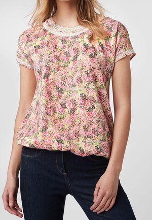 Woman wearing a short-sleeve floral print blouse with lace trim on the neckline and sleeve edges, paired with dark blue jeans.