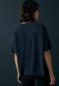 Námořnická modrá oversized tričko vyrobené z měkké bavlny, s krátkými rukávy a kulatým výstřihem, viděné zezadu na tmavém pozadí.