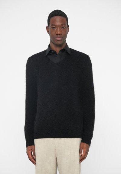 Schwarzer, flauschiger V-Ausschnitt-Pullover aus weichem Material, kombiniert mit einem schwarzen Hemd und hellbeigen Hosen, vor einem einfarbigen Hintergrund stehend.