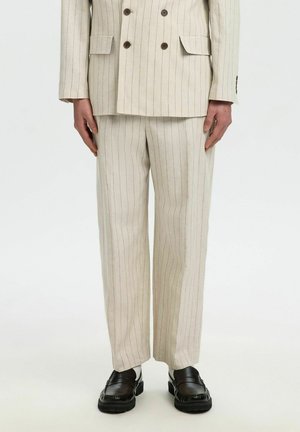 Homme portant un pantalon de costume beige à fines rayures et un blazer assorti, associé à des mocassins en cuir noir et des chaussettes blanches, debout face à l'avant.
