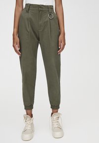 Pantalon fuselé vert olive en tissu léger, avec un détail de bague argentée à la taille et des poignets élastiques. Porté avec des baskets beiges.