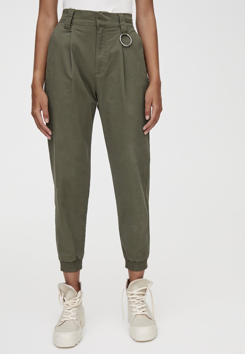 Pantalon fuselé vert olive en tissu léger, avec un détail de bague argentée à la taille et des poignets élastiques. Porté avec des baskets beiges.