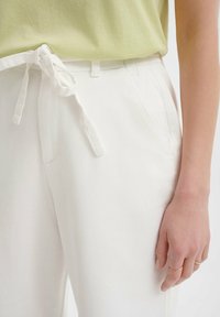 Pantalones blancos con una textura suave y una cinta de tela en la cintura, combinados con una blusa verde claro. Bolsillos visibles en los lados de los pantalones.
