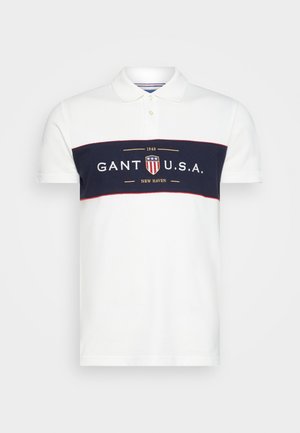Polo blanc avec une rayure horizontale bleu marine, arborant le logo "GANT U.S.A." en or. Patte de boutonnage à trois boutons et manches courtes.