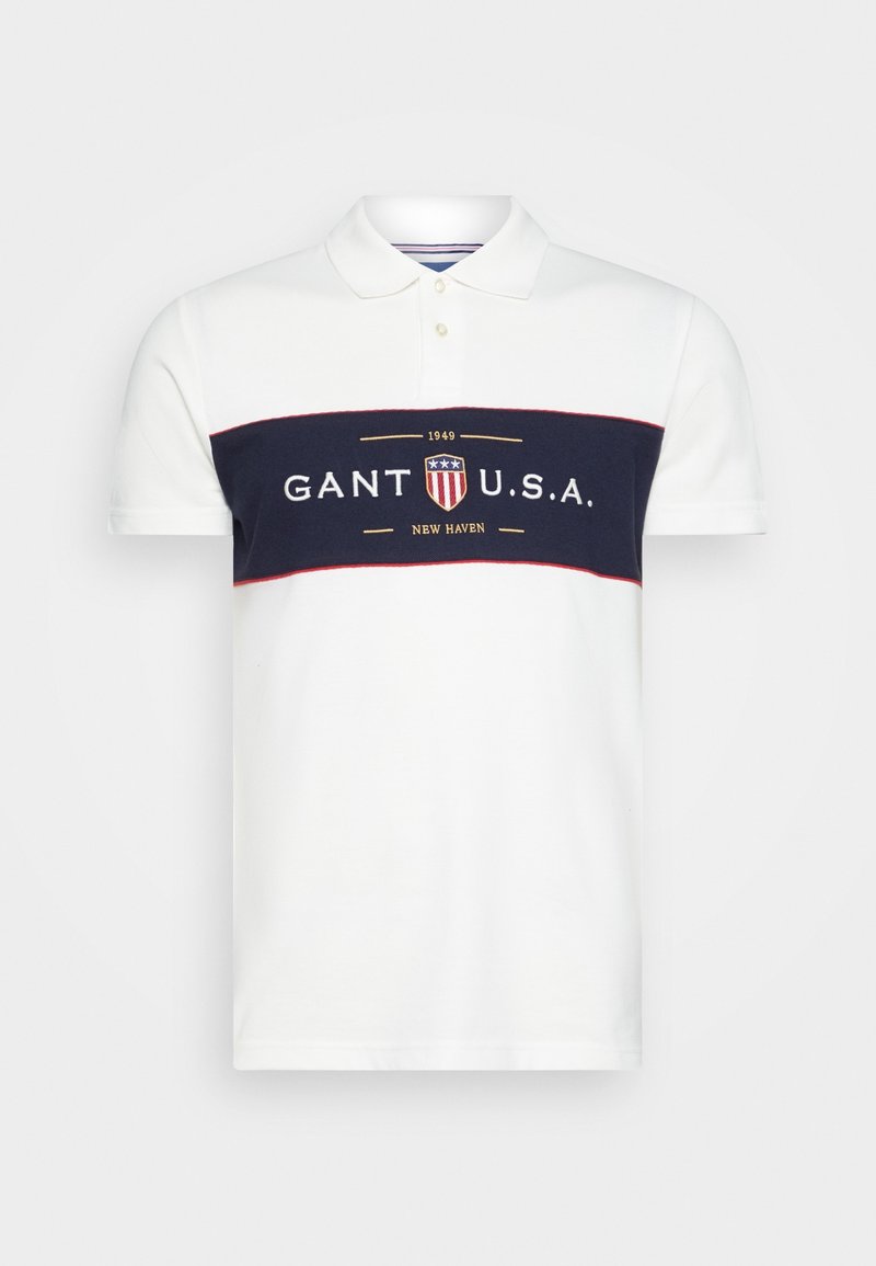 Polo blanc avec une rayure horizontale bleu marine, arborant le logo "GANT U.S.A." en or. Patte de boutonnage à trois boutons et manches courtes.