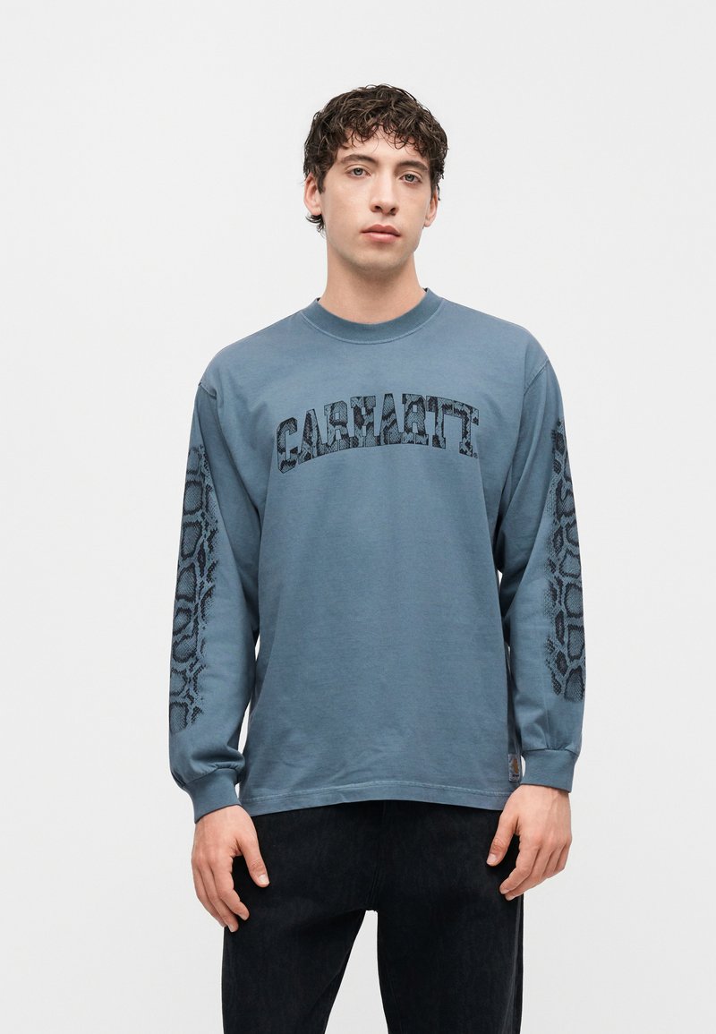 Νέος άνδρας φοράει μπλε μακρυμάνικο πουκάμισο Carhartt με λεπτομέρειες φιδιού στα μανίκια, στέκεται μπροστά σε λευκό φόντο.