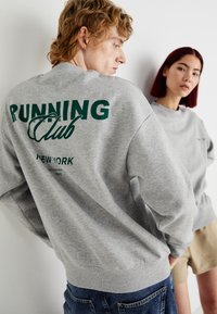 Grijze sweatshirt met "RUNNING Club" en "NEW YORK" in het groen gedrukt. Casual fit met geribbelde manchetten, gedragen met denim shorts door een model.