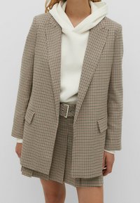 Blazer à carreaux en tissu léger aux tons beige et marron, assorti d'un sweat à capuche blanc et d'un short à carreaux assorti. Doté d'une ceinture en guise d'accent.
