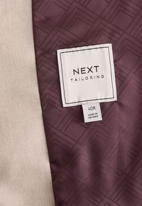 Étiquette de vêtement « NEXT TAILORING » cousue sur un tissu bordeaux avec un motif géométrique subtil, à côté d’un tissu beige texturé. Taille 40R, fabriqué au Vietnam.