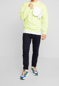 Sudadera verde neón, camisa blanca en capas, pantalones oscuros y zapatillas coloridas con acentos azules, verdes y naranjas; pequeña bolsa blanca.