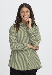 Giovane donna che sorride con la mano vicino al mento, indossa una camicia a righe verticali verdi e bianche e pantaloni neri.